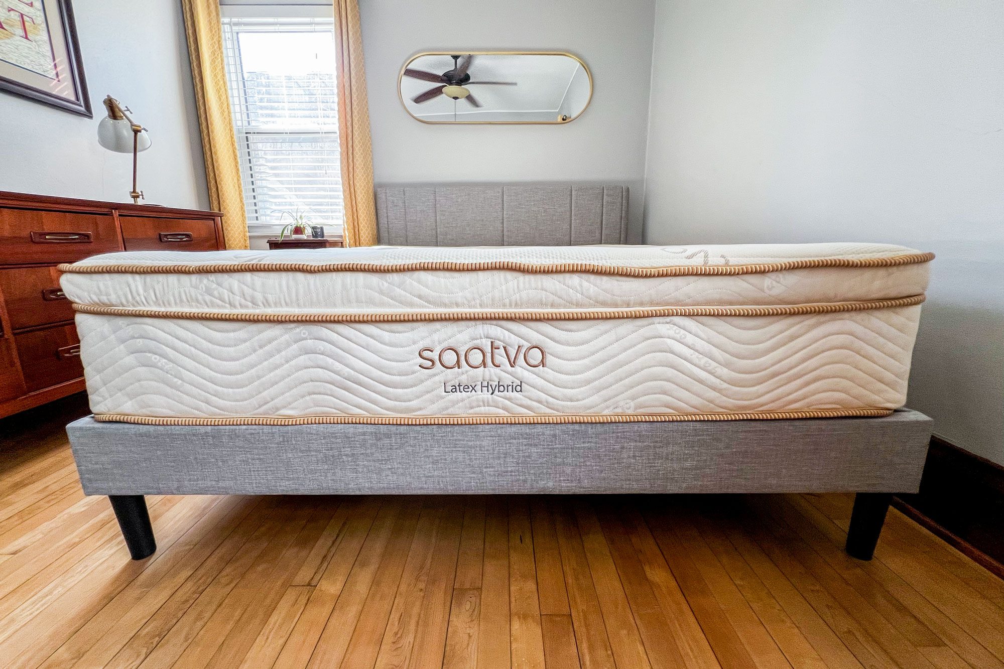 Mattress resting on a bed frame, labeled "saatva Latex Hybrid," in a bedroom with wood flooring, a dresser, and a window with curtains.