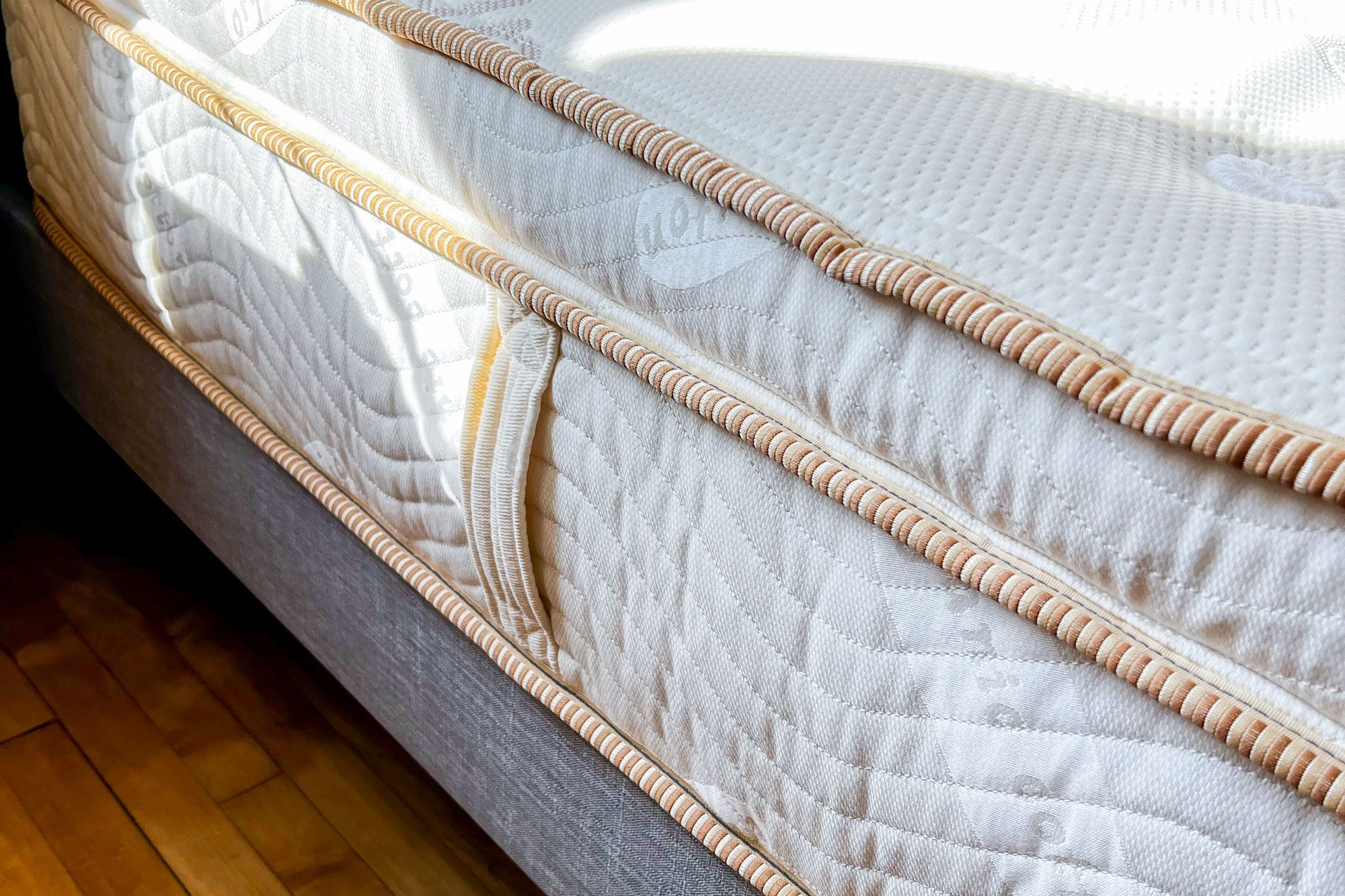 Mattress displayed with stitched handle, set atop a wooden floor. Sunlight softly illuminates the textured surface and ornate edging.
