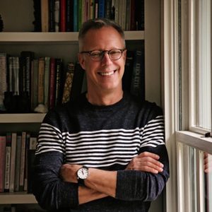 A person smiles while standing by a window, wearing glasses and a striped sweater, with arms crossed. Books fill shelves in the background.