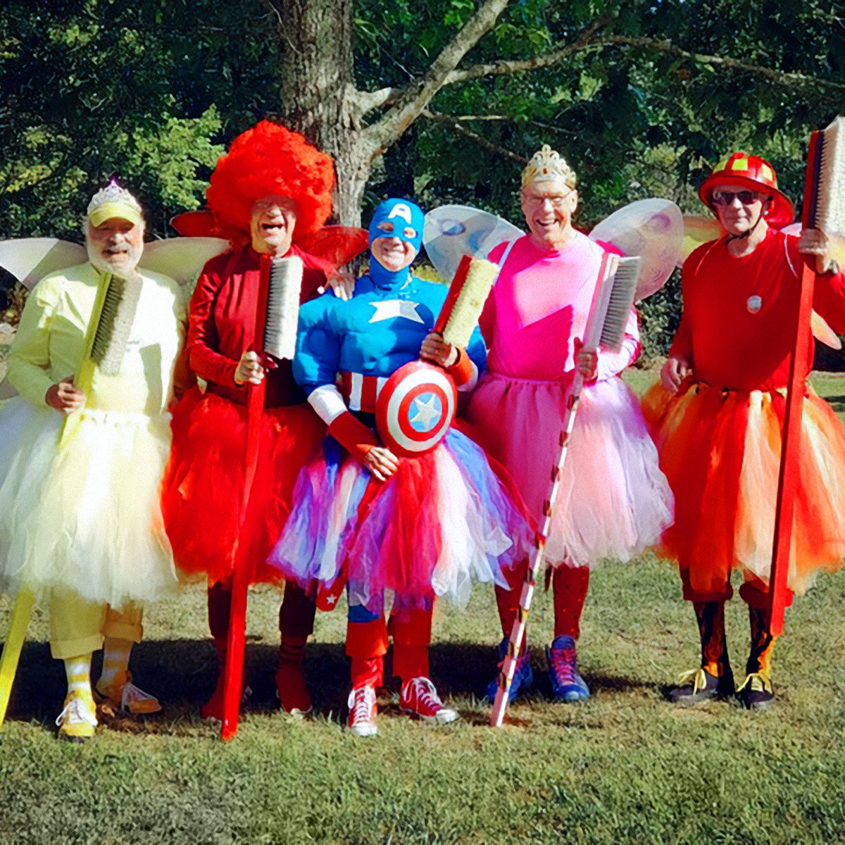 Five people wear colorful costumes and tutus, holding oversized brushes, standing on grass with trees in the background, smiling and posing together.