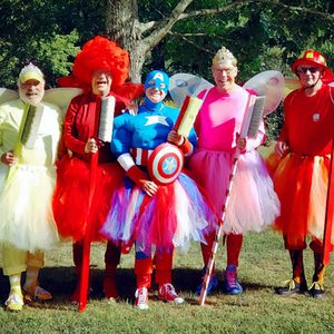 Five adults wearing colorful costumes hold brooms, posing in a grassy, park-like setting near trees.