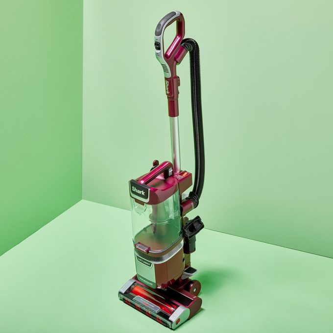 Vacuum cleaner standing idle on a mint-green surface, featuring a transparent dust container and red accents, against a light green wall backdrop.