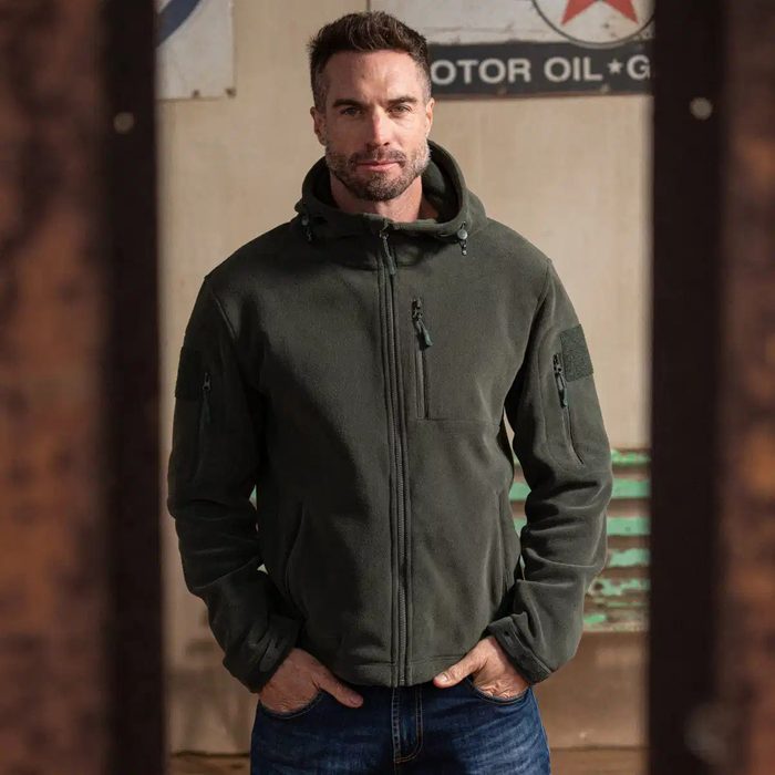 Man stands with hands in pockets, wearing a green jacket, in a rustic indoor setting with vintage signs on the walls.
