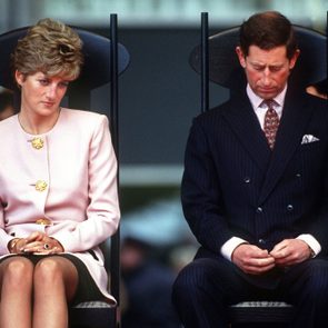 Two people sit on chairs, appearing thoughtful and reserved, in a formal outdoor setting with a blurred crowd in the background.