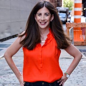 A person stands smiling, wearing a bright orange top, hands on hips, on a cobblestone street with construction materials nearby.