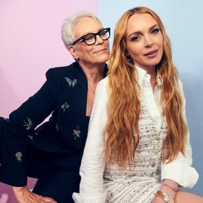 Two people pose together, smiling, in front of pastel pink and blue backgrounds. One wears glasses and a dark outfit, the other a white dress.