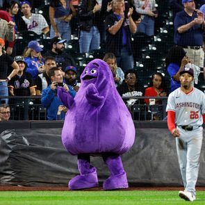 Purple mascot stands clapping, surrounded by a cheering crowd at a baseball game with a player walking past in a Washington uniform.