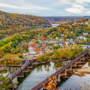 Rail bridge crosses river; vibrant autumn leaves surround a picturesque town nestled in a lush valley with rolling hills.