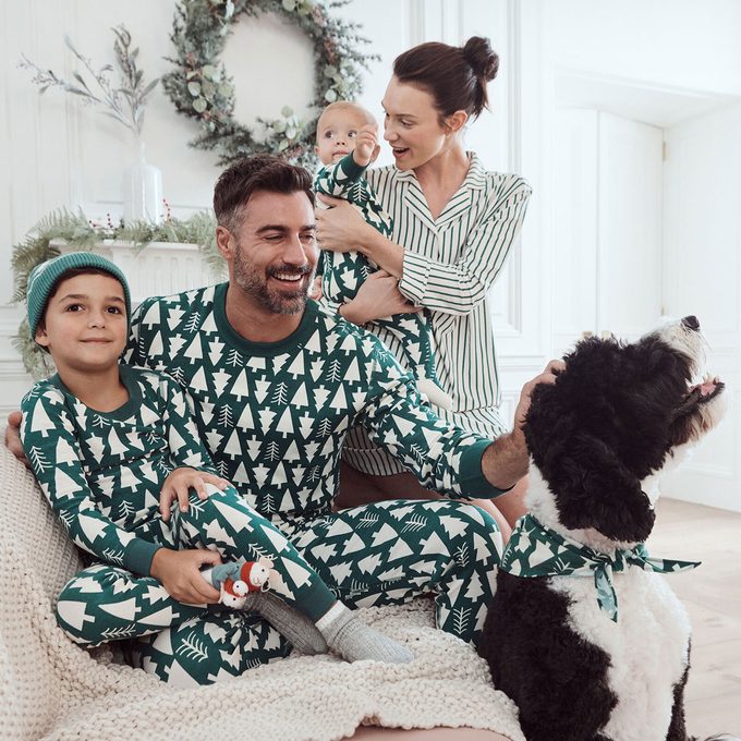 Family wearing matching tree-patterned pajamas smiles while sitting indoors, petting a large black-and-white dog, with holiday wreaths and dÃ©cor around.