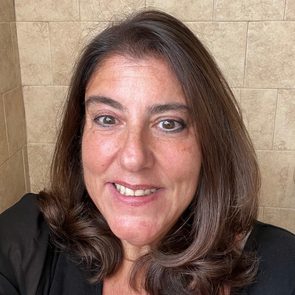 A person smiles widely in front of a beige tiled wall, creating a warm and friendly atmosphere.