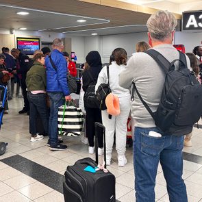 People wait at an airport gate, carrying bags and luggage, under a sign displaying 