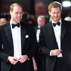 Two men in tuxedos walking and smiling, surrounded by a crowd on a red carpet event with bright lights in the background.