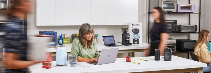 A person works on a laptop in a kitchen appliance testing area, with shelves of appliances around and others moving past.