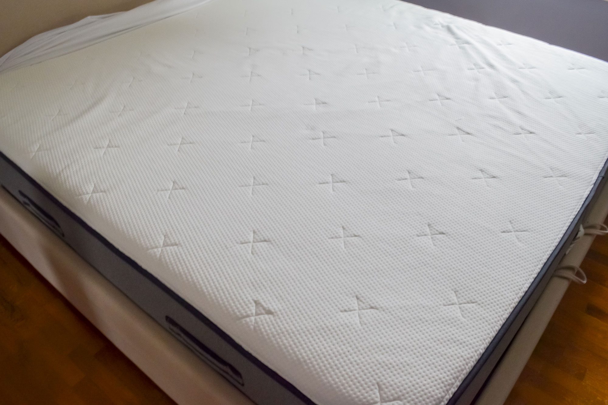 A large white mattress with textured cross patterns sits on a wooden floor in a sparse room.
