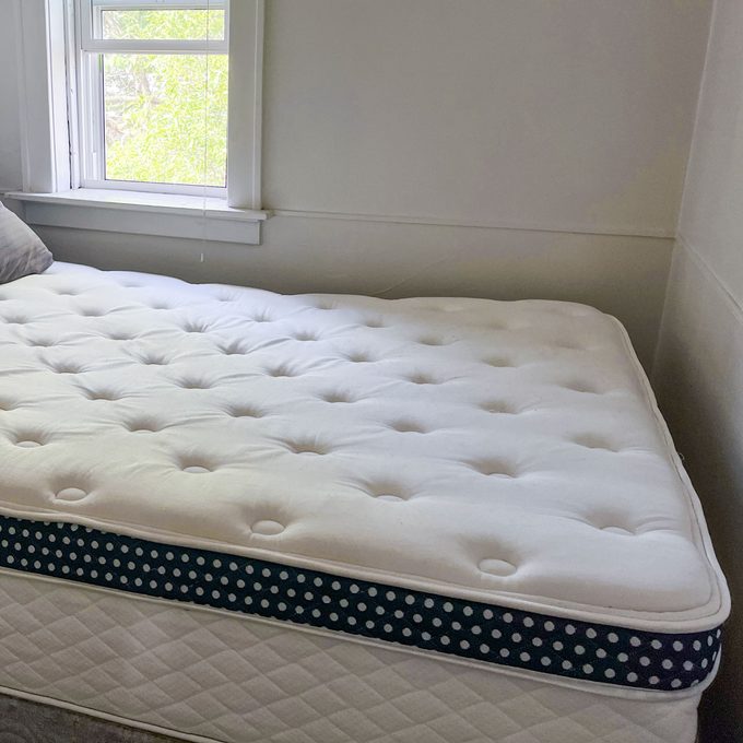 Mattress lies on bed frame, featuring tufted white surface and blue polka dot side. Room is softly lit with a window nearby.