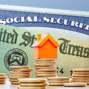 Orange house rests on stacked coins, surrounded by more coins, against a backdrop of U.S. Social Security and Treasury documents.