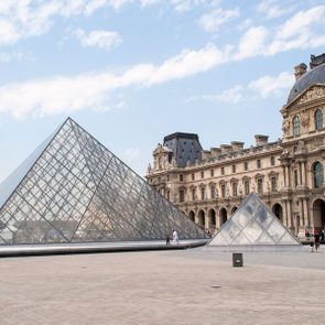 Glass pyramid stands still, surrounded by classic architecture and visitors, under a blue sky with scattered clouds.