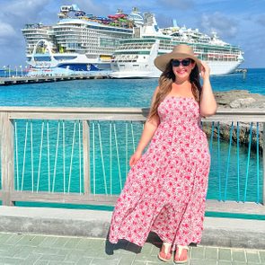 Woman poses by railing, holding her hat, wearing a floral dress. Background features docked cruise ships and clear blue sea under a sunny sky.