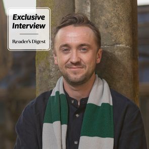 Man smiling, wearing a striped scarf, leaning against a stone wall. Text reads: 