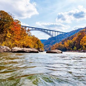 Bridge arches over a flowing river surrounded by vibrant autumn trees under a partly cloudy sky.