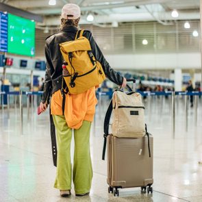 Traveler pulling suitcase, wearing a yellow backpack and bright clothing, walks through an airport terminal with flight information displays overhead.