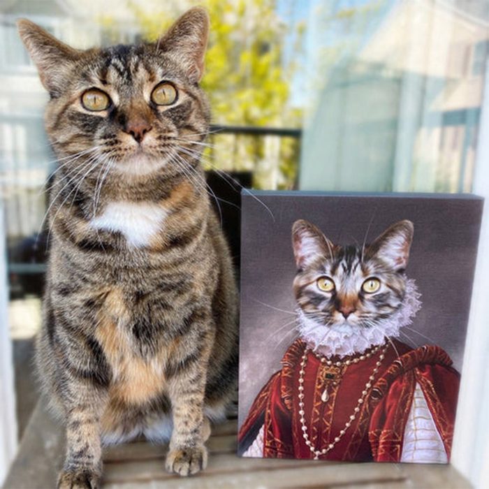 Tabby cat sits next to a painted portrait of itself in royal attire, outdoors on a sunny day.