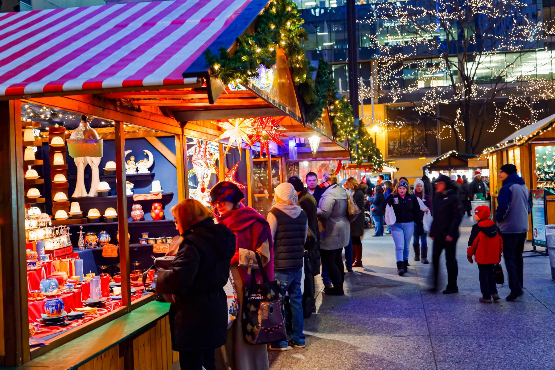Market stalls display festive goods; people browse under decorative lights. Trees are adorned with twinkling lights in a lively outdoor setting.