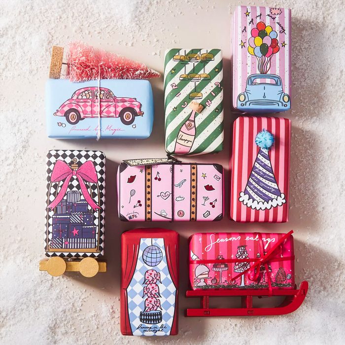 Colorful wrapped gifts are artistically arranged on a snowy surface, each decorated with unique festive illustrations and themes, including cars, hats, and champagne bottles.