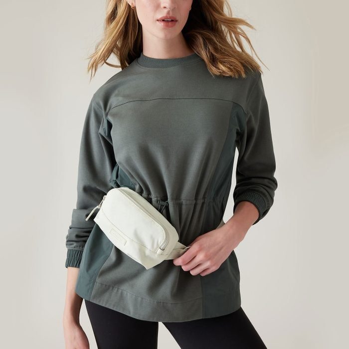 A person stands wearing a green pullover and black pants, holding a white waist bag, against a plain light background.