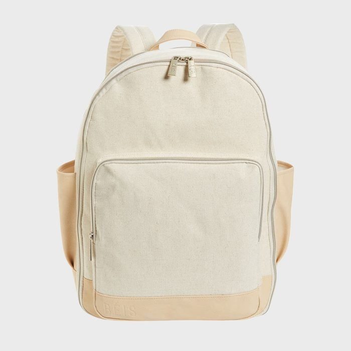 A cream-colored backpack with tan accents is standing upright; it has a front pocket and beige straps, set against a plain gray background.