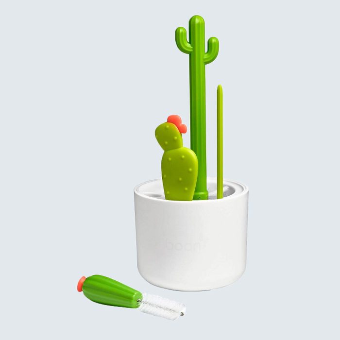 Cactus-shaped bottle brushes stand upright in a white holder; one brush with a flower lies nearby, against a light background.