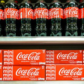 Coca-Cola bottles and mini can packs displayed on a grocery shelf, showcasing the brand's logo and original taste label.