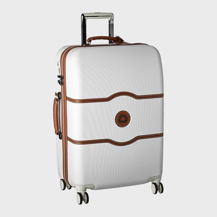 Suitcase stands upright with extended handle; white exterior features brown leather accents; four wheels provide mobility; against a plain gray background.