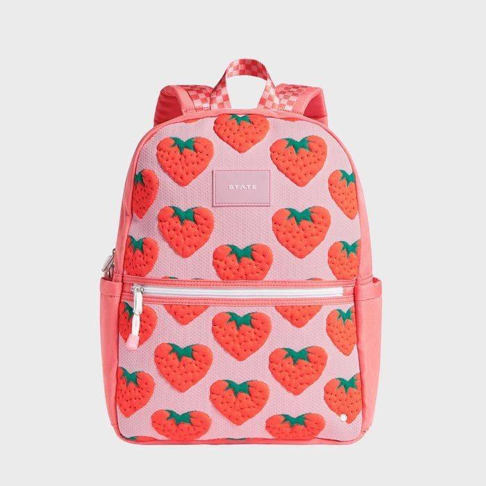A backpack displays strawberry patterns; it rests upright against a gray background. The label 