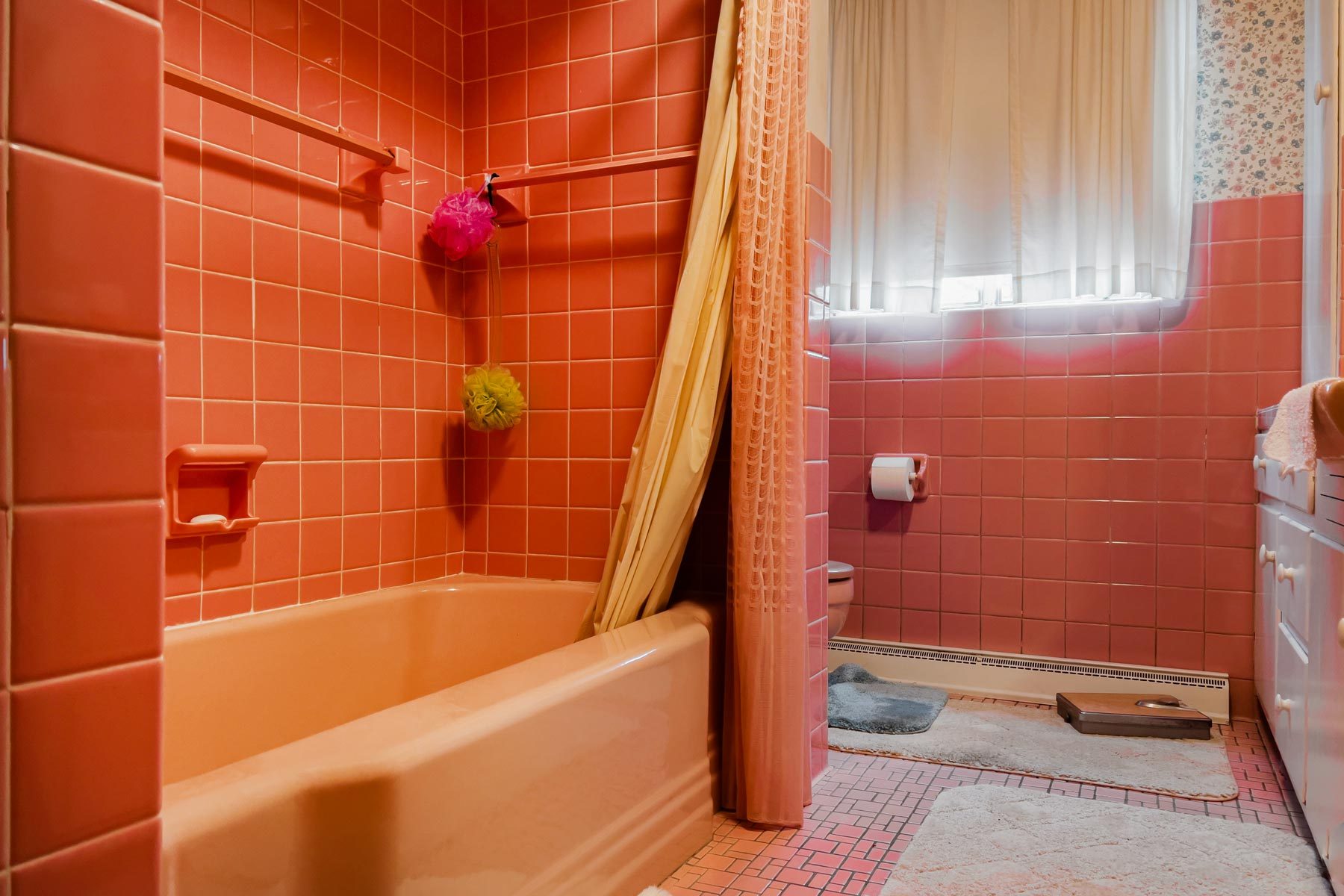 Pink-tiled bathtub situated with shower curtain, colorful sponges, in a vintage bathroom featuring a toilet, rugs, and a window with curtains.
