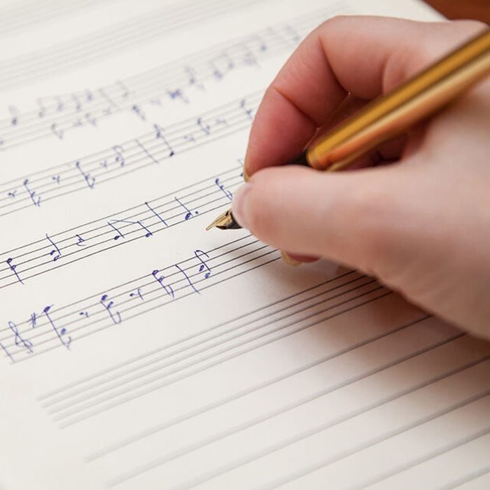 A hand writes musical notes with a gold pen on sheet music paper in a bright setting.