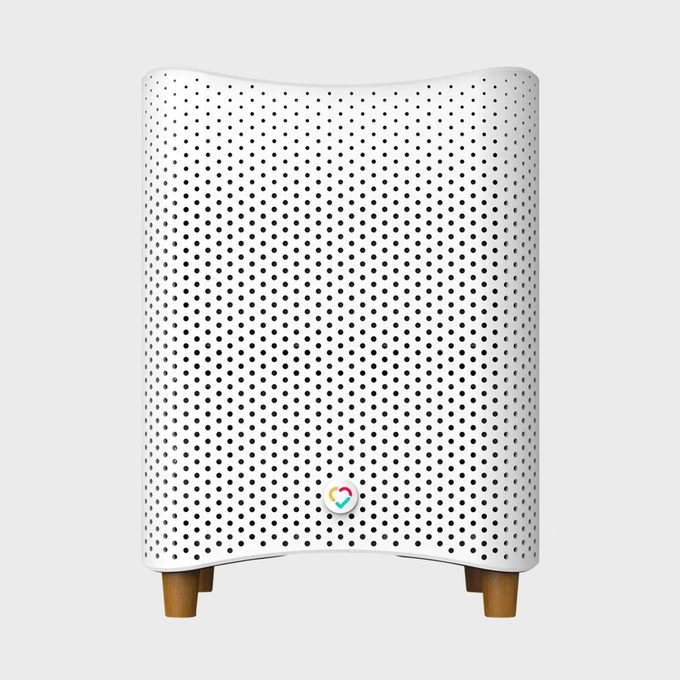White air purifier, standing on wooden legs, featuring a perforated design and a colorful heart-shaped logo in a minimalistic setting.