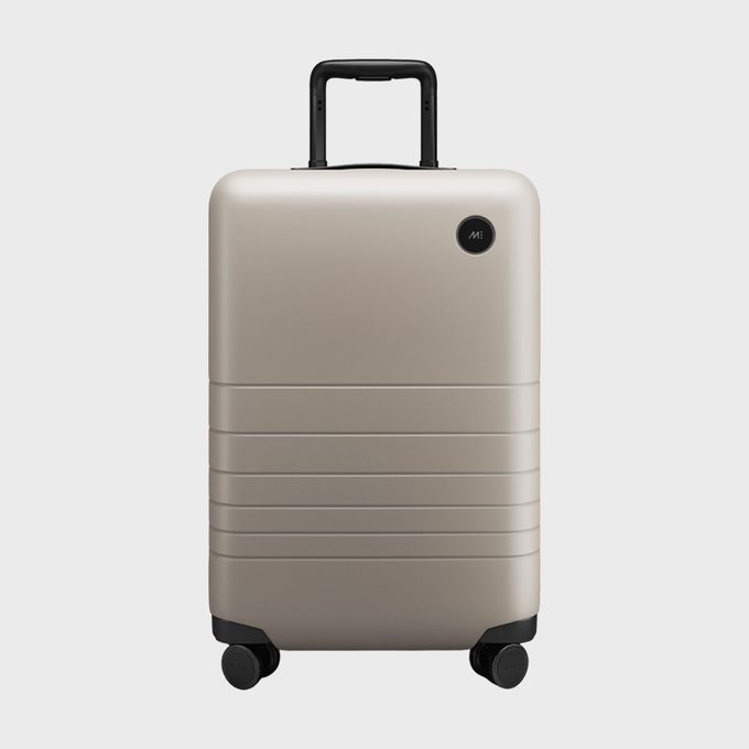 Beige suitcase stands upright; has black handle and four wheels; smooth, minimal design with horizontal grooves; plain white background.
