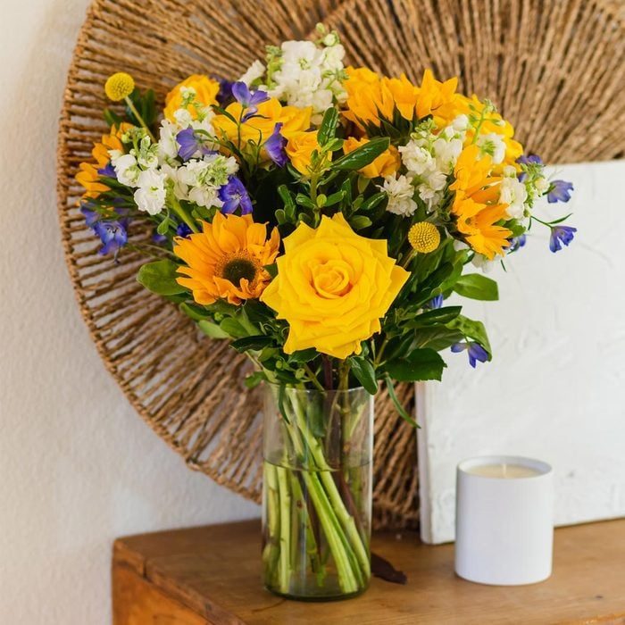 Bouquet with yellow sunflowers, seated in a glass vase, on a wooden table beside a white candle, framed by a woven wall decoration.