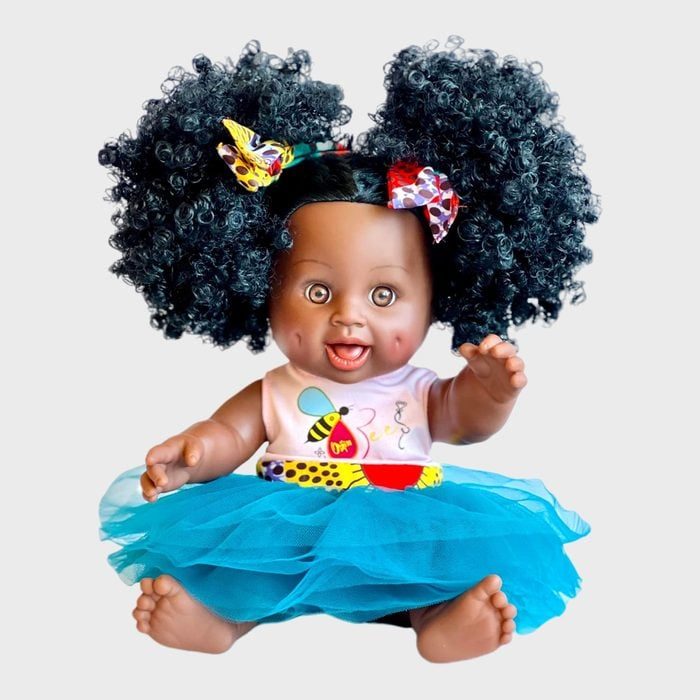 Doll with curly hair, smiling playfully, wearing a colorful dress and blue tutu; bows adorn the hair, set against a plain backdrop.