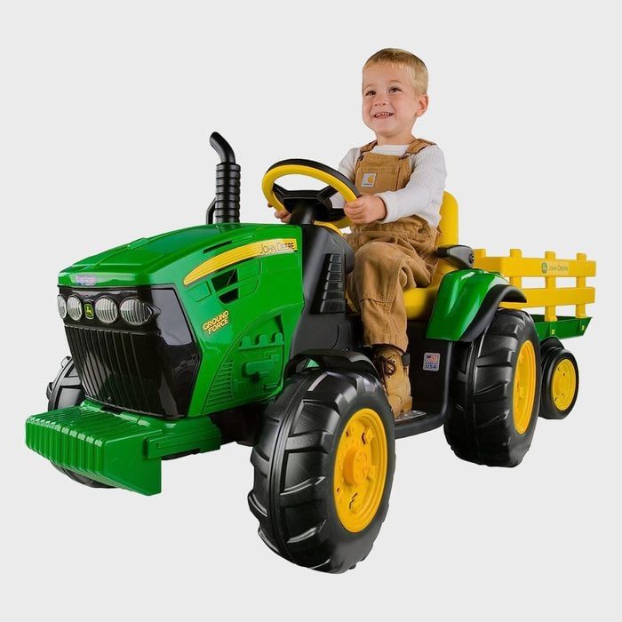 Child driving a green toy tractor with a yellow trailer, indoors on a plain background.