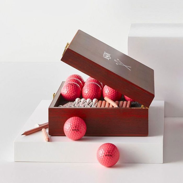 Wooden box holds golf balls and cigars, lid open, resting on white blocks; environment is minimalistic with no visible text.