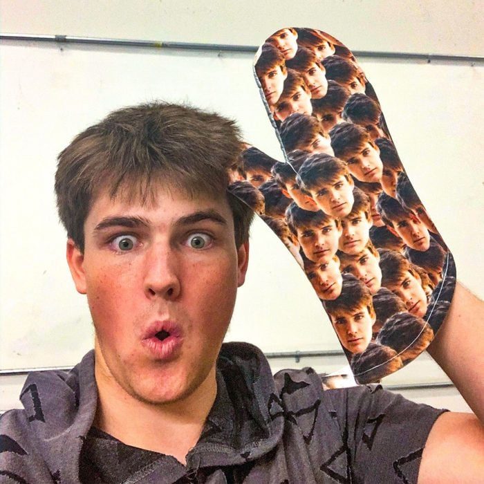 Person holding oven mitt printed with many faces, making surprised expression in a room with plain walls.