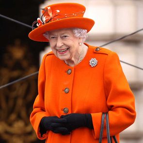 Elderly woman smiling, wearing an orange coat and hat, black gloves, pearl necklace, and brooch; standing outside with blurred architectural background.