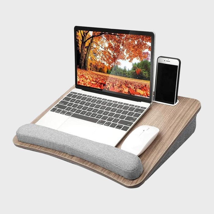 Laptop resting on a wooden lap desk displaying an autumn scene; accompanied by a phone in a slot and a mouse beside a gray wrist pad.