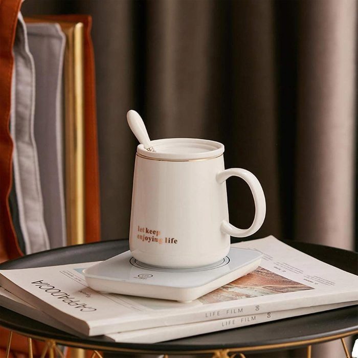 A white mug with a lid and spoon rests on a warmer, set atop books with 