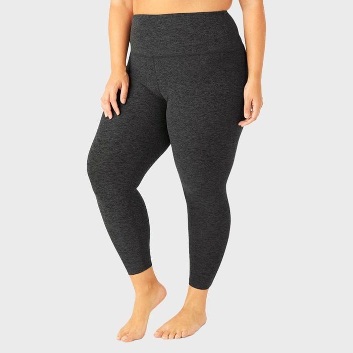 Barefoot person in dark leggings stands against a plain gray background, showing the leggings' fit and high waistband.
