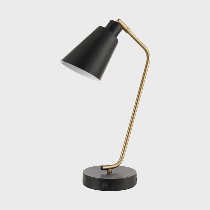 A black lamp with a cone shade and gold arm, standing on a black circular base with a power switch, set against a plain backdrop.