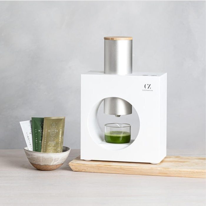 Machine dispenses liquid into a glass; nearby, packets sit in a bowl. The setting is a minimal countertop with light background.