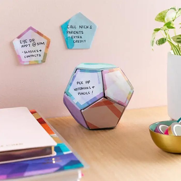 Colorful dodecahedron displays reminders, sitting on a desk with other stationery items and a plant nearby. Two notes are pinned on the wall.